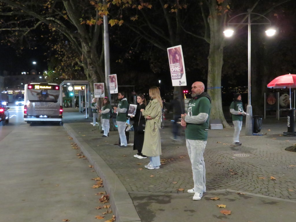 Aktivist*innen stehen auf dem Trottoir und halten verschiedene A2 Schilder mit der Aufschrift "Stoppt Primatenversuche!" oder "Für Forschung ohne Primatenversuche!" sowie "LSCV" in den Händen. Am Boden liegen weitere Transparente mit denselben Motiven, rundherum sind einzelne Passant*innen. Alle Aktivist*innen tragen einen weissen Schutzanzug und ein grünes LSCV-Shirt.