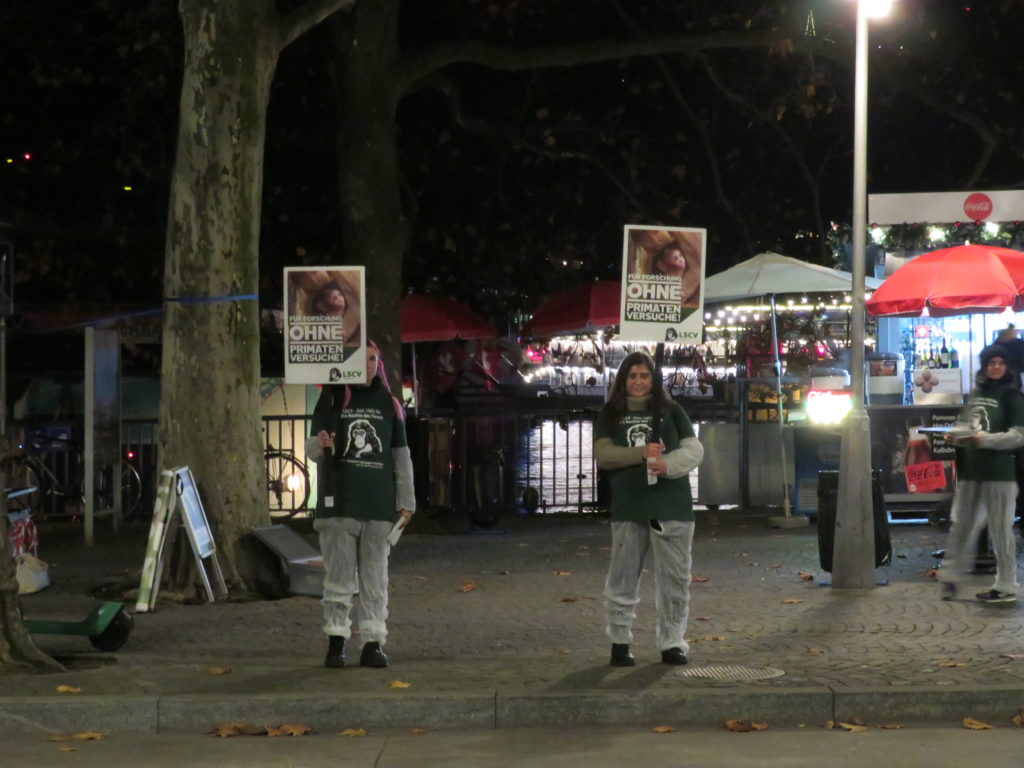 Aktivist*innen stehen auf dem Trottoir und halten verschiedene A2 Schilder mit der Aufschrift "Stoppt Primatenversuche!" oder "Für Forschung ohne Primatenversuche!" sowie "LSCV" in den Händen. Am Boden liegen weitere Transparente mit denselben Motiven, rundherum sind einzelne Passant*innen. Alle Aktivist*innen tragen einen weissen Schutzanzug und ein grünes LSCV-Shirt.