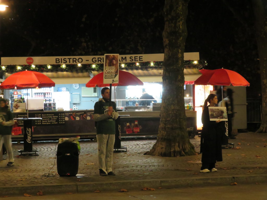 Aktivist*innen stehen auf dem Trottoir und halten verschiedene A2 Schilder mit der Aufschrift "Stoppt Primatenversuche!" oder "Für Forschung ohne Primatenversuche!" sowie "LSCV" in den Händen. Am Boden liegen weitere Transparente mit denselben Motiven, rundherum sind einzelne Passant*innen. Alle Aktivist*innen tragen einen weissen Schutzanzug und ein grünes LSCV-Shirt.