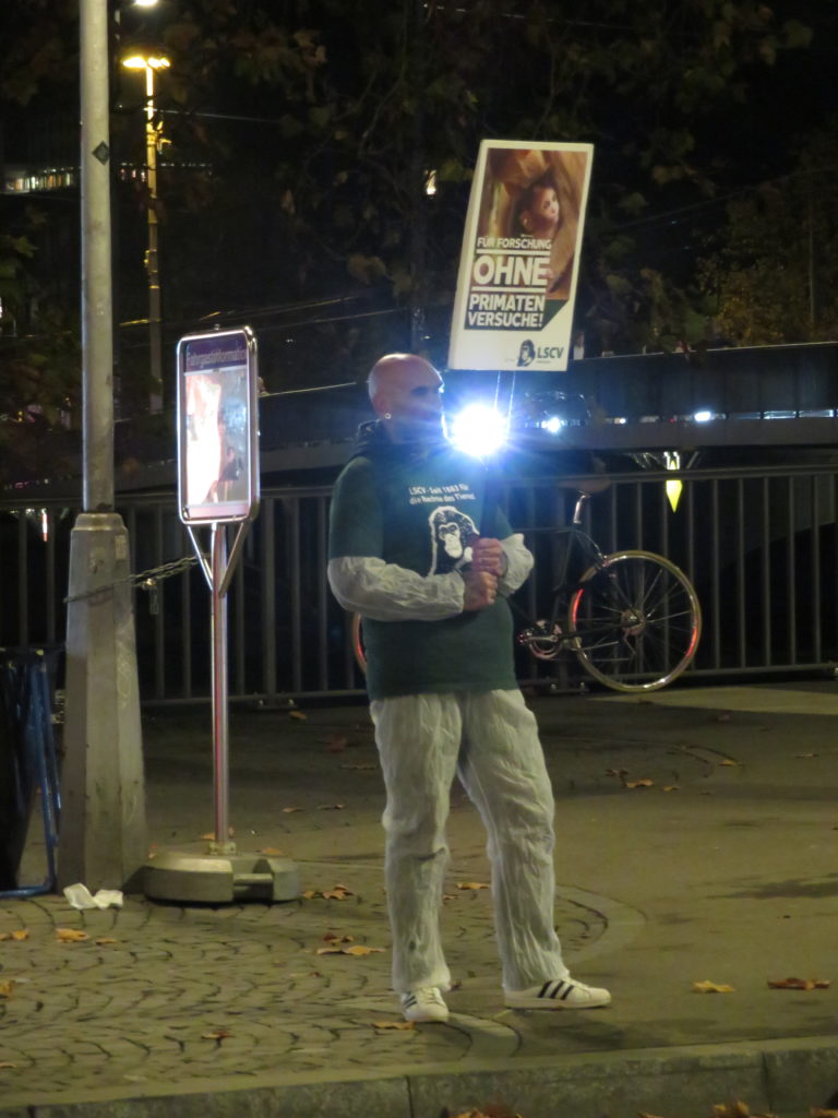 Ein Aktivist steht auf dem Trottoir und hält ein A2 Schild mit der Aufschrift "Für Forschung ohne Primatenversuche!" sowie "LSCV" in den Händen. Am Boden liegen weitere Transparente mit denselben Motiven, rundherum sind einzelne Passant*innen. Alle Aktivist*innen tragen einen weissen Schutzanzug und ein grünes LSCV-Shirt.