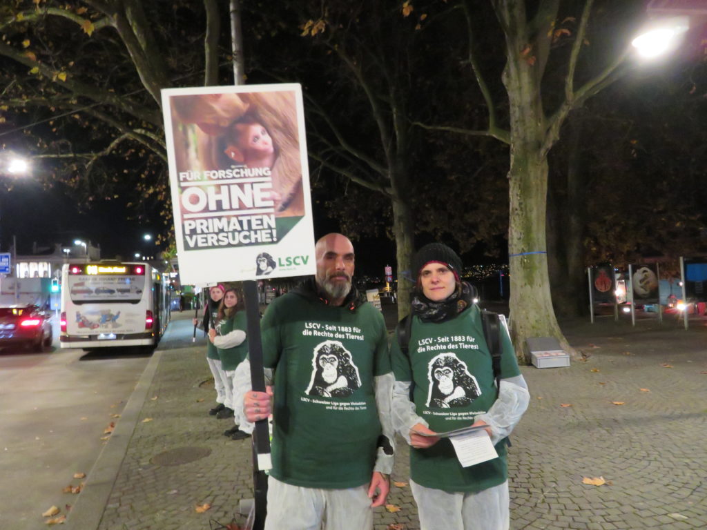 Zwei Aktivist*innen schauen in die Kamera während sie auf dem Trottoir stehen und halten ein A2 Schild mit der Aufschrift "Für Forschung ohne Primatenversuche!" sowie "LSCV" in den Händen. Am Boden liegen weitere Transparente mit denselben Motiven, rundherum sind einzelne Passant*innen. Alle Aktivist*innen tragen einen weissen Schutzanzug und ein grünes LSCV-Shirt.