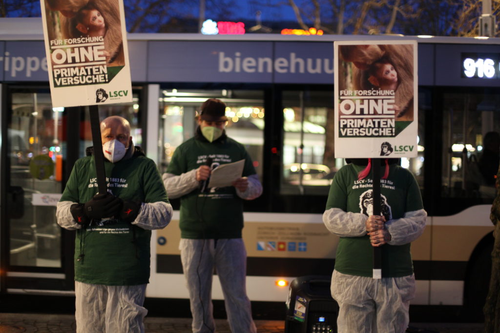 Zwei Aktivist*innen stehen auf dem Trottoir und halten A2 Schilder mit der Aufschrift "Für Forschung ohne Primatenversuche!" sowie "LSCV" in den Händen. Dahinter steht ein*e Aktivist*in und hält eine Rede. Alle Aktivist*innen tragen einen weissen Schutzanzug, ein grünes LSCV-Shirt und eine FFP2-Maske.