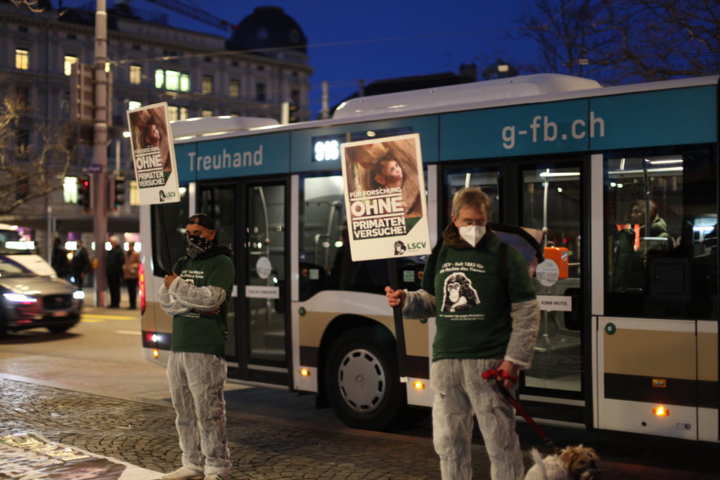 Zwei Aktivist*innen stehen auf dem Trottoir und halten A2 Schilder mit der Aufschrift "Für Forschung ohne Primatenversuche!" sowie "LSCV" in den Händen. Ein Aktivist hat einen Hund an der Leine. Am Boden liegen weitere Transparente mit denselben Motiven. Alle Aktivist*innen tragen einen weissen Schutzanzug, ein grünes LSCV-Shirt und eine Mund-Nasen-Abdeckung.