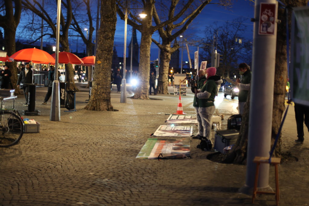 Aktivist*innen stehen auf dem Trottoir und halten A2 Schilder. Daneben ist sind Stehtische und Passant*innen zu sehen. Am Boden liegen weitere Transparente mit denselben Motiven. Alle Aktivist*innen tragen einen weissen Schutzanzug, ein grünes LSCV-Shirt und eine Mund-Nasen-Abdeckung.