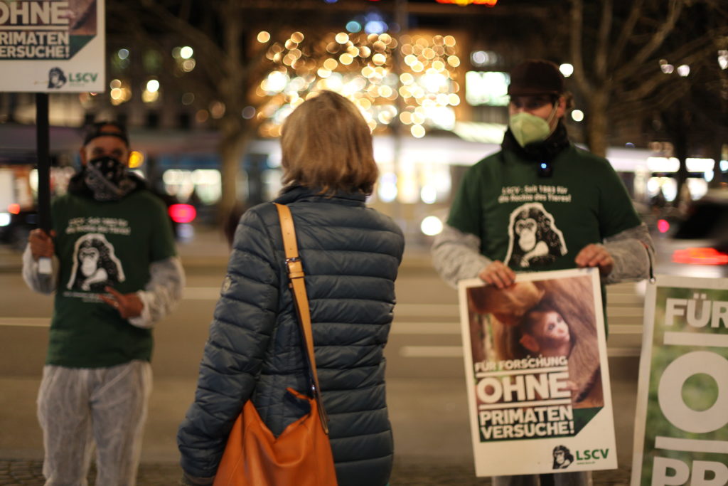 Zwei Aktivist*innen reden mit eine*r Passant*in. Beide Aktivist*innen tragen einen weissen Schutzanzug, ein grünes LSCV-Shirt und eine Mund-Nasen-Abdeckung.