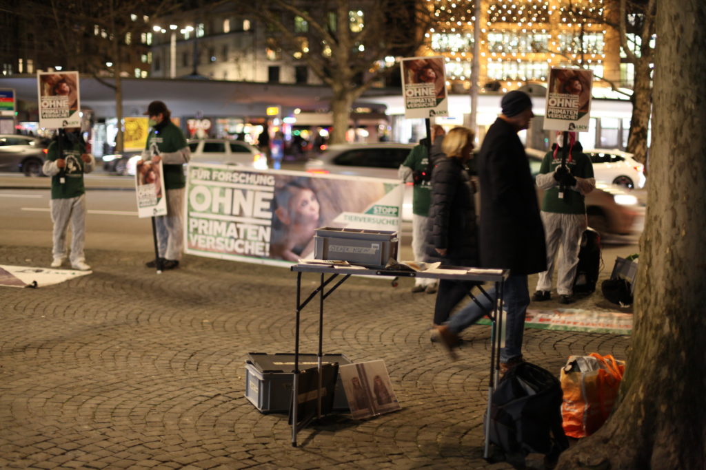 Die Aktion von weiter weg zu sehen. Im Vordergrund der Materialtisch mit Unterschriftenlisten, Aufklebern und Flyern. Dahinder Halten Aktivist*innen ein Transparent mit der Aufschrift "Für Forschung ohne Primatenversuche!" sowie "Stopp Tierversuche" in den Händen. Links und rechts weitere Aktivist*innen mit Schildern. Alle Aktivist*innen tragen einen weissen Schutzanzug, ein grünes LSCV-Shirt und eine Mund-Nasen-Abdeckung.